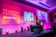 The host of the Museum + Heritage Awards stands at a lectern on a stage. The Museum + Heritage Awards logo is on the wall behind her